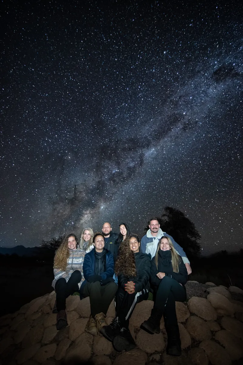 Grupo de 7 viajeros sentados sobre piedras contemplando la Vía Láctea
