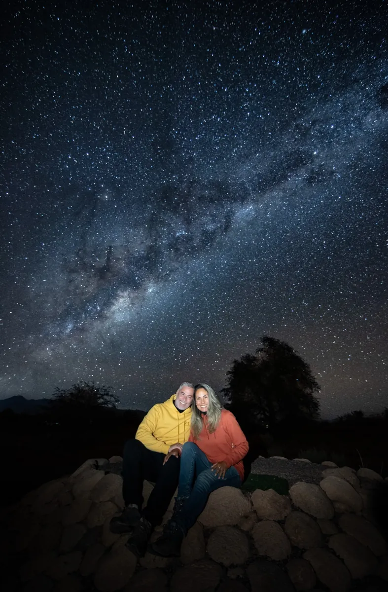 Pareja iluminada con luz naranja y amarilla sonriendo bajo la Vía Láctea