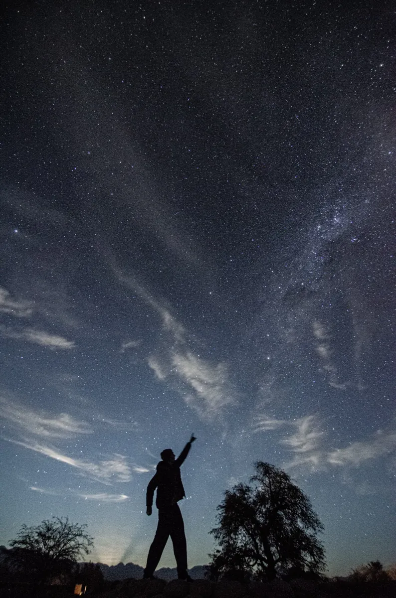 Silueta de persona señalando el cielo bajo nubes y Vía Láctea en tonos azules